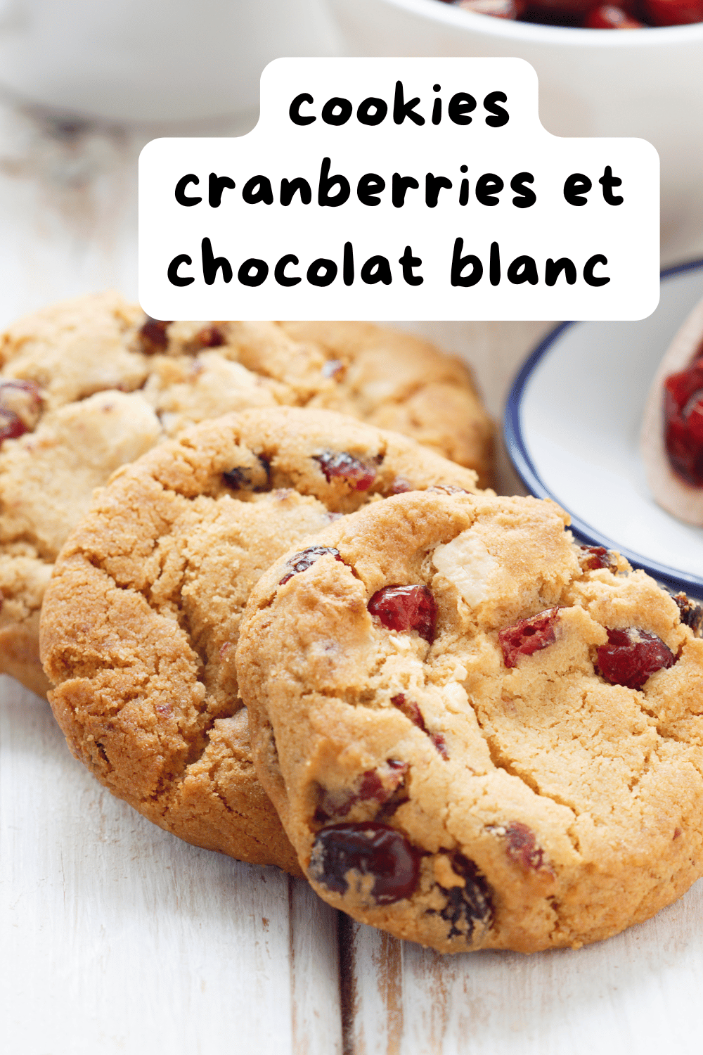 Cookies cranberries et chocolat blanc dorés, disposés sur une grille de refroidissement avec des morceaux de chocolat blanc et des cranberries en arrière-plan.