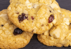 Cookies cranberries et chocolat blanc dorés, disposés sur une grille de refroidissement avec des morceaux de chocolat blanc et des cranberries en arrière-plan.