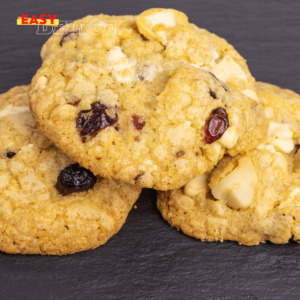 Cookies cranberries et chocolat blanc dorés, disposés sur une grille de refroidissement avec des morceaux de chocolat blanc et des cranberries en arrière-plan.