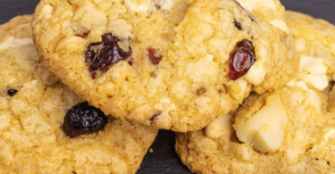 Cookies cranberries et chocolat blanc dorés, disposés sur une grille de refroidissement avec des morceaux de chocolat blanc et des cranberries en arrière-plan.