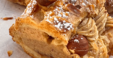 Un Paris-Brest doré, garni d’une crème mousseline pralinée, saupoudré de sucre glace et décoré de noisettes.