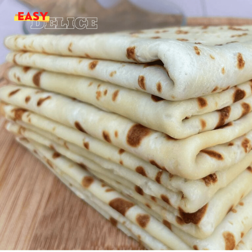 Une pile de crêpes maison dorées, garnies de Nutella et de fruits, sur une table avec un fouet et une casserole à côté.