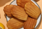 Madeleines dorées et moelleuses, légèrement bombées, parfumées au citron, servies sur une assiette élégante avec des zestes de citron.