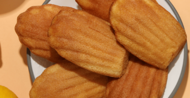 Madeleines dorées et moelleuses, légèrement bombées, parfumées au citron, servies sur une assiette élégante avec des zestes de citron.